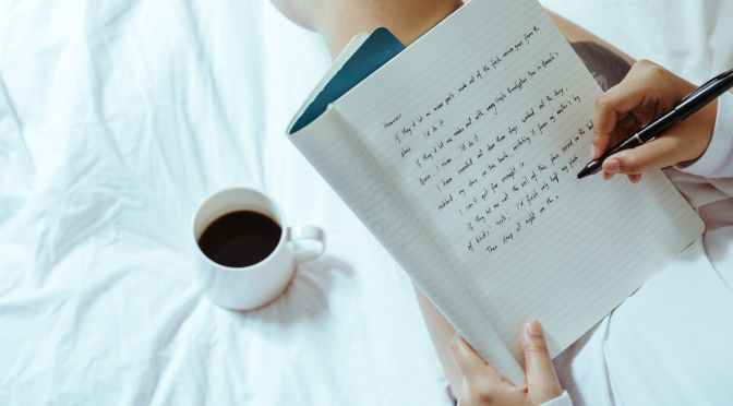 woman with coffee writing in notebook on bed